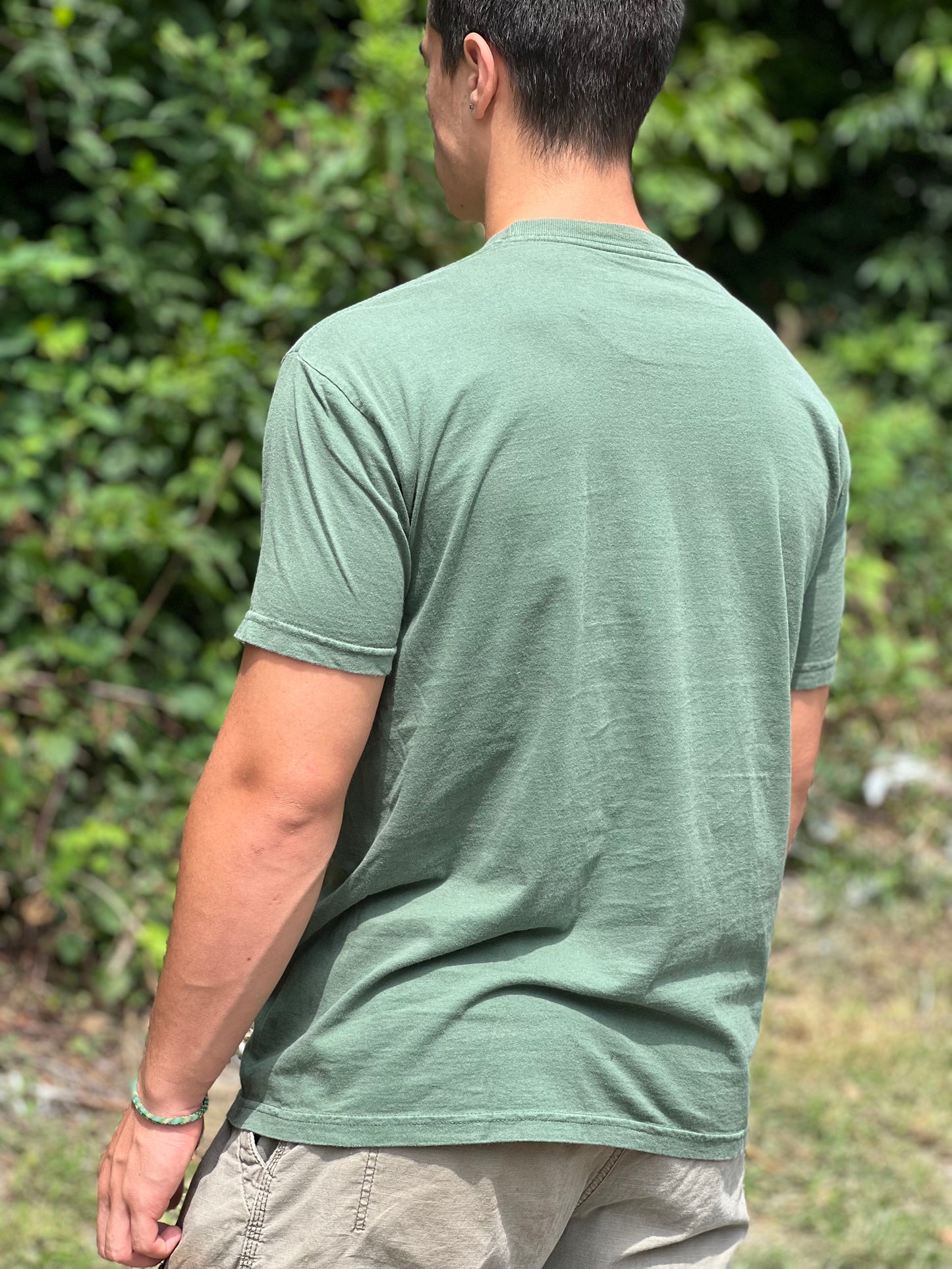 Person wearing a green t-shirt standing outdoors with greenery in the background