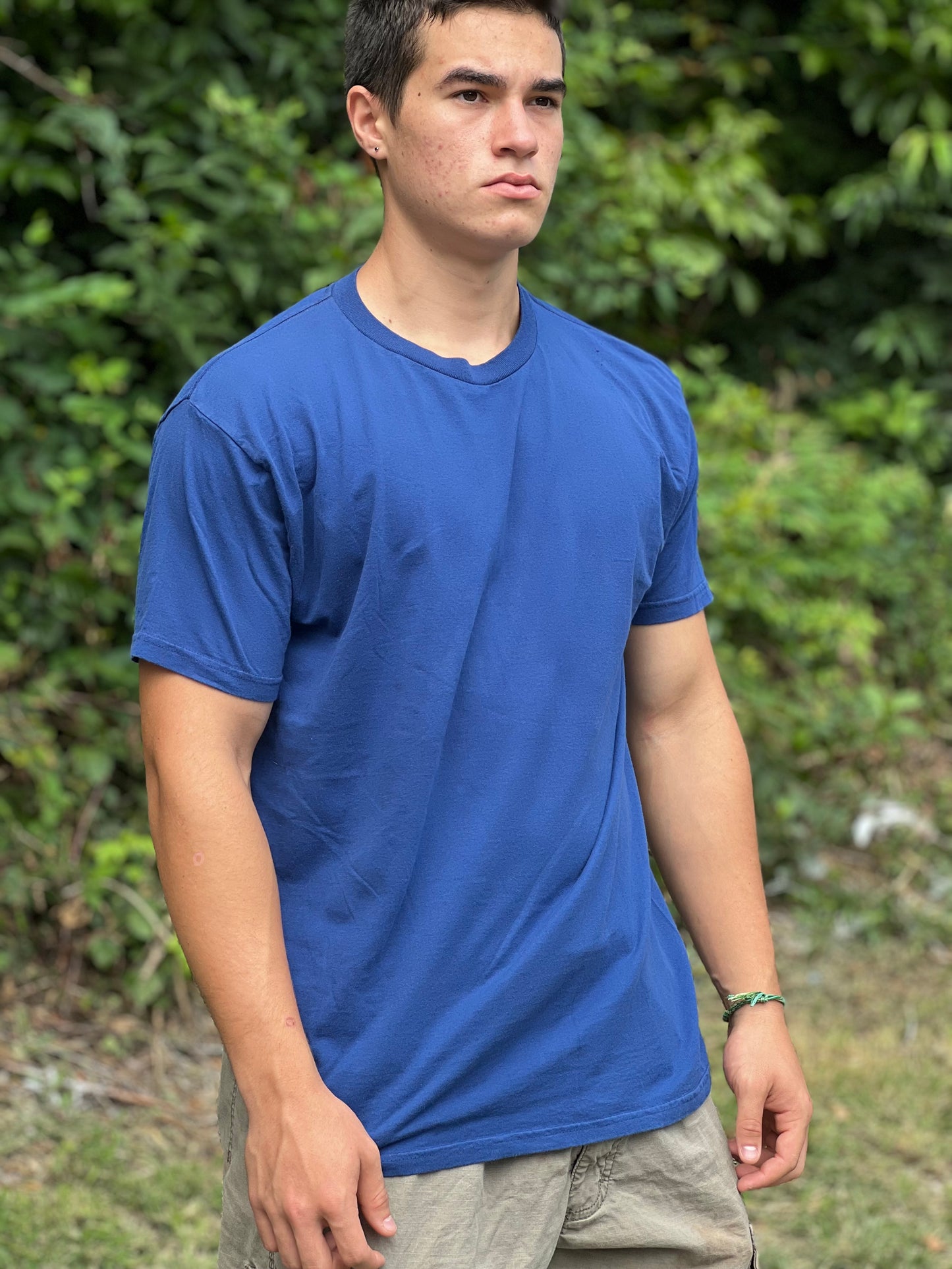Person wearing a blue t-shirt standing outdoors with greenery in the background