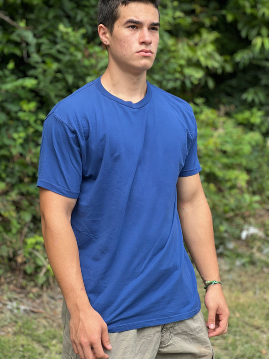 Person wearing a blue t-shirt standing outdoors with greenery in the background