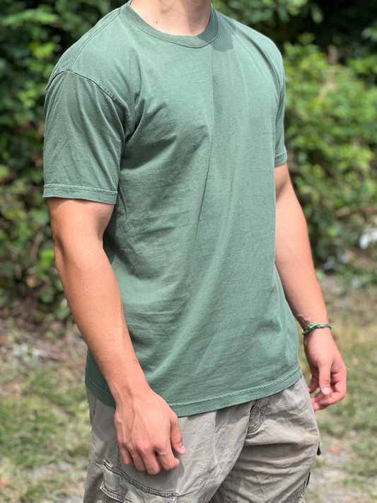 Person wearing a green t-shirt and gray shorts standing outdoors with greenery in the background